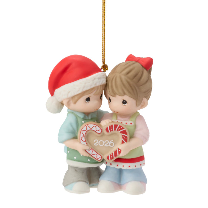 A Precious Moments-style Christmas ornament featuring a couple figurine holding a heart-shaped candy cane. The boy wears a red Santa hat and light blue sweater, and the girl wears a red bow and green apron.