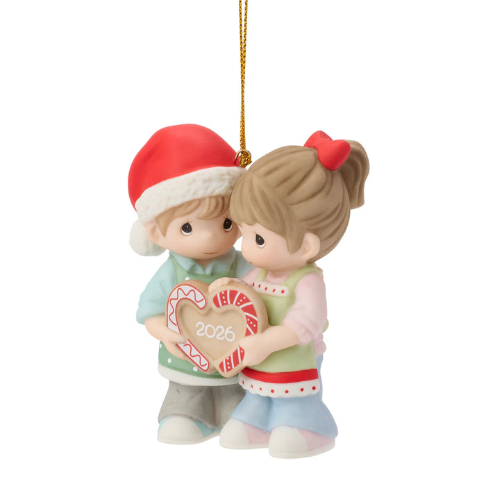 A Precious Moments Christmas ornament depicting a young boy in a red Santa hat and a young girl with a red bow holding a heart-shaped candy cane with '2026' written on it, suspended by a gold string against a white background.
