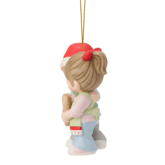 A ceramic Christmas ornament depicting a mother and child embracing, shown from behind. The ornament features a red Santa hat, light brown hair, a green vest, and light blue pants, suspended by a gold string against a plain white background.