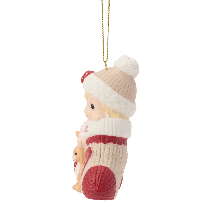 A Christmas ornament shaped like a baby doll wearing a cream knit hat with a white pom-pom and a red-and-cream sweater, holding a small dog, hanging from a gold string on a white background.