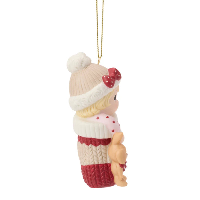 A Christmas ornament featuring a baby doll dressed in a cream knit hat with a red bow, a cream sweater with red hearts, and a red knit skirt, holding a small teddy bear. The ornament hangs from a gold string against a plain white background.