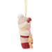 A Christmas ornament featuring a baby doll dressed in a cream knit hat with a red bow, a cream sweater with red hearts, and a red knit skirt, holding a small teddy bear. The ornament hangs from a gold string against a plain white background.