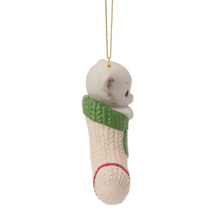 A Christmas ornament shaped like a baby bear peeking out of a knitted cream stocking with a green cuff, hanging from a gold string against a white background.