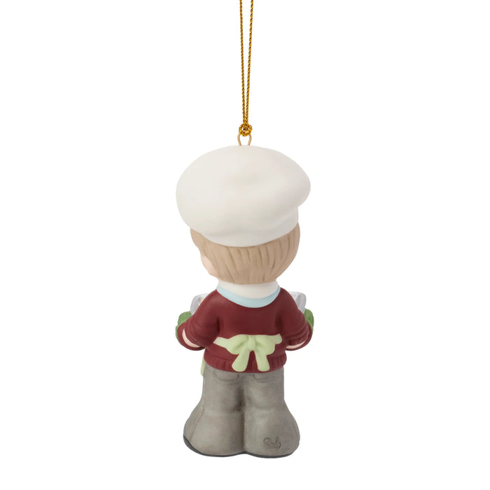 A Christmas ornament shaped like a child dressed as a chef, shown from the back. The figure has a white hat, a burgundy sweater with a pale green bow, gray pants, and a gold hanging string against a plain white background.