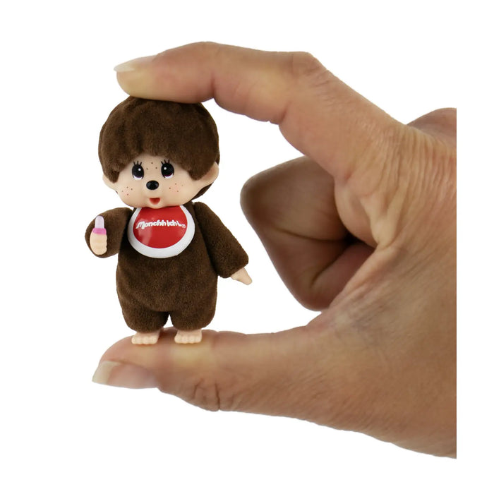 A small brown plush monkey toy with a red-and-white chest emblem, shown being held between a person's fingers against a plain white background.