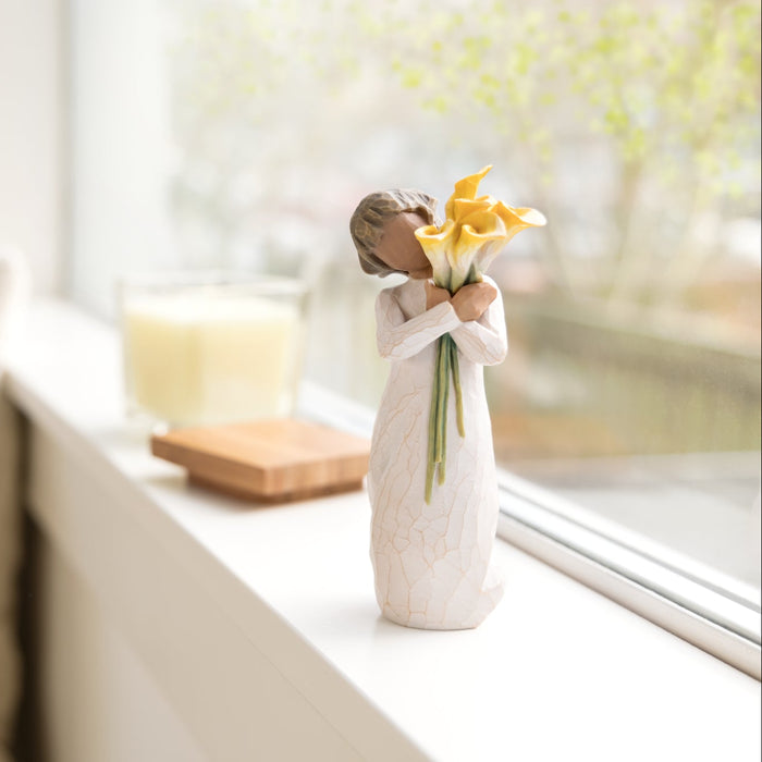 Standing figure in cream dress with darker skin tone and hair color, holding large bouquet of yellow calla lilies in arms
