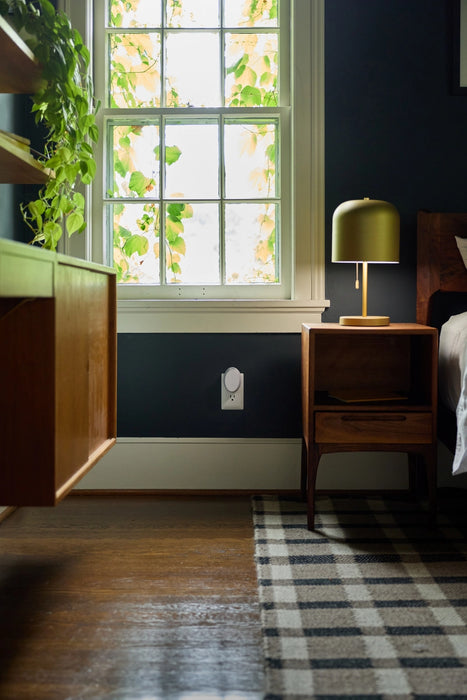 A styled bedroom corner featuring a dark navy blue wall, a wooden nightstand with a green dome lamp, a window with ivy outside, and a black and white plaid rug on a dark wood floor.