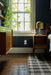 A styled bedroom corner featuring a dark navy blue wall, a wooden nightstand with a green dome lamp, a window with ivy outside, and a black and white plaid rug on a dark wood floor.