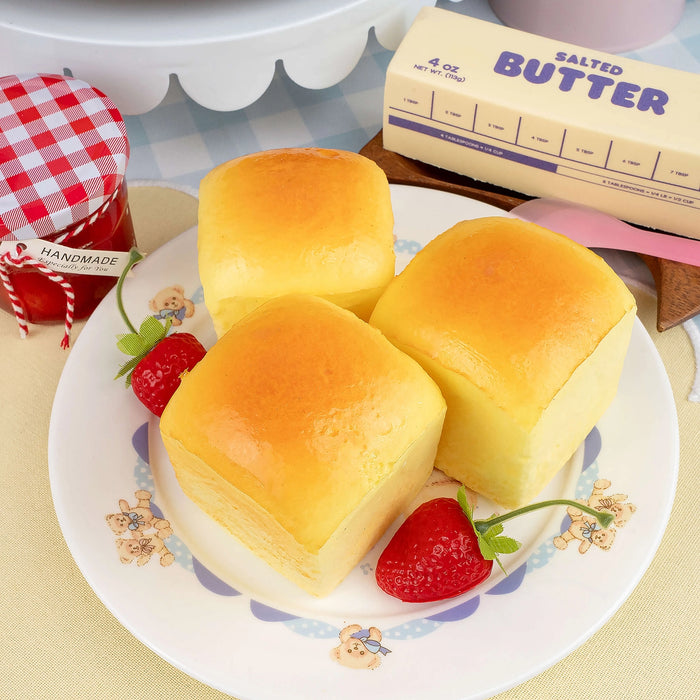Three bread rolls on a decorative plate with strawberries and a stick of salted butter in the background.