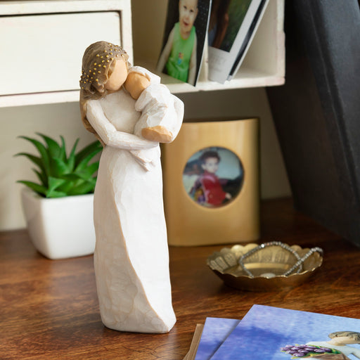 Statue of a woman holding a child on a wooden surface with photos and a plant in the background.