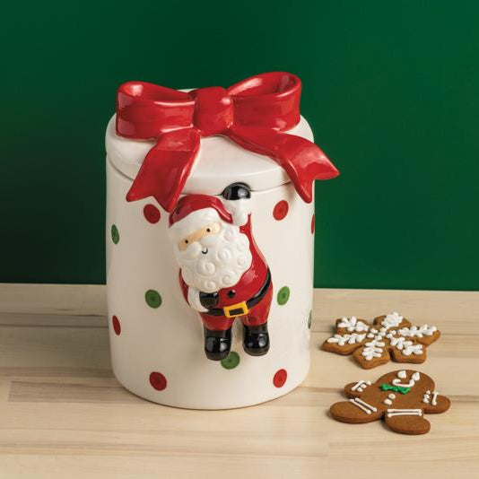 Ceramic cookie jar with Santa design and red bow on a wooden surface with cookies.