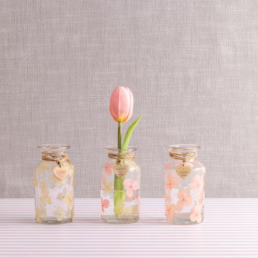 Three small glass jars with floral designs containing a single pink tulip on a textured gray background. 