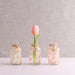 Three small glass jars with floral designs containing a single pink tulip on a textured gray background. 