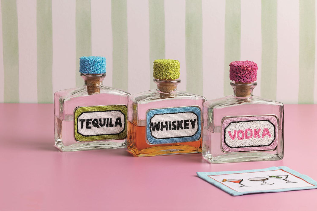 Three decorative bottles labeled 'Tequila', 'Whiskey', and 'Vodka' on a pink surface with a striped background.