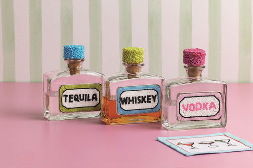 Three decorative bottles labeled 'Tequila', 'Whiskey', and 'Vodka' on a pink surface with a striped background.
