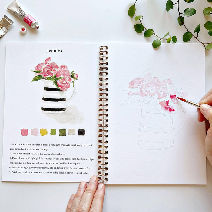 Open art book with a hand holding a paintbrush, surrounded by greenery and art supplies.