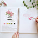 Open art book with a hand holding a paintbrush, surrounded by greenery and art supplies.