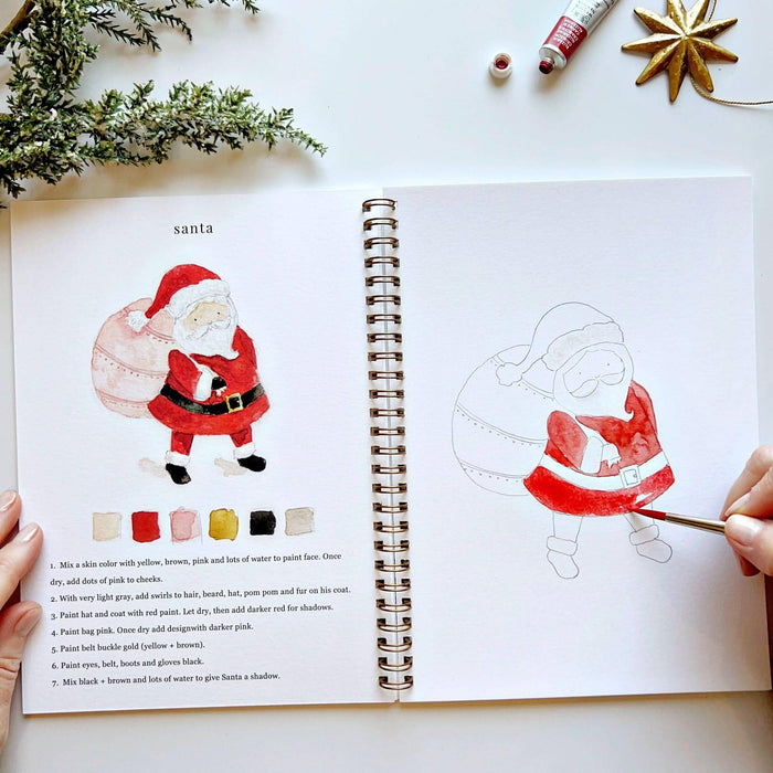 Open book with a drawing of Santa Claus and paint swatches, surrounded by Christmas decorations.