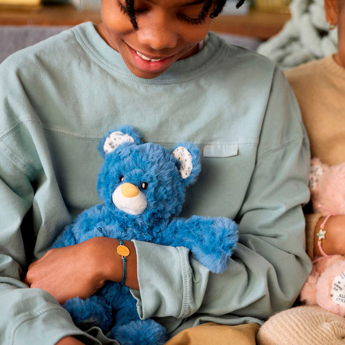 Person holding a blue teddy bear with another person partially visible in the background.