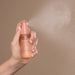 Hand holding a pink spray bottle labeled 'Akitsch' with mist being released against a beige background.