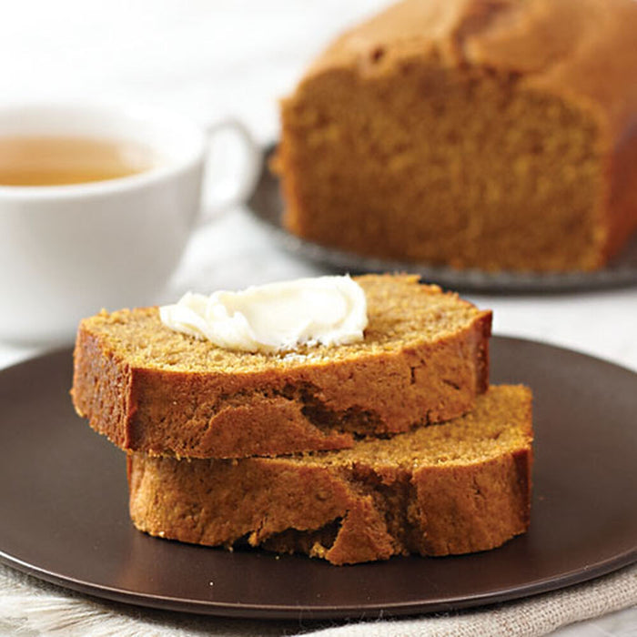 Two slices of brown bread with butter on a dark plate, with a loaf of bread and a cup of tea in the background.