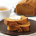 Two slices of brown bread with butter on a dark plate, with a loaf of bread and a cup of tea in the background.