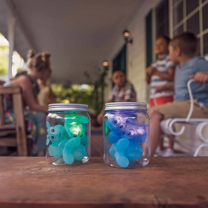 Two jars with glowing blue and green firefly-like creatures on a wooden table, with people in the background.