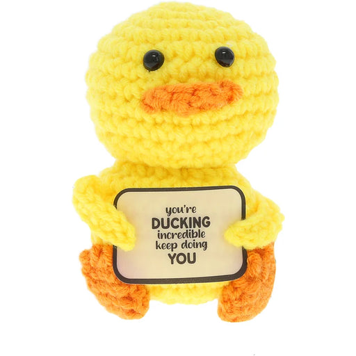 A yellow crocheted duck plush toy holding a cream-colored sign that reads 'you're DUCKING incredible keep doing YOU'. The toy has black bead eyes and orange accents on its beak and feet, set against a plain white background.