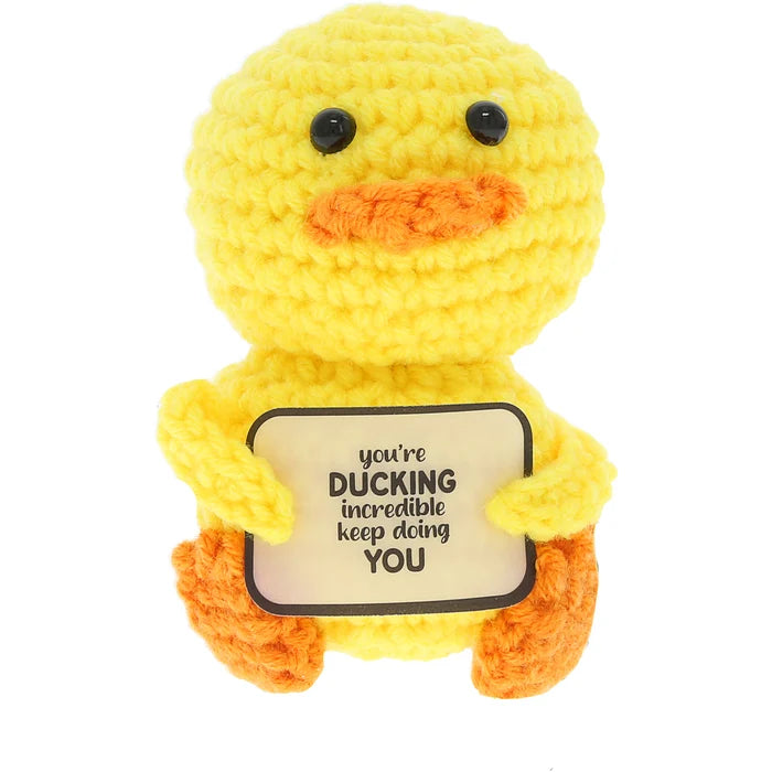 A yellow crocheted duck plush toy holding a cream-colored sign that reads 'you're DUCKING incredible keep doing YOU'. The toy has black bead eyes and orange accents on its beak and feet, set against a plain white background.