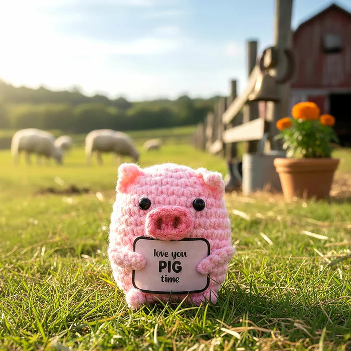 A pink crocheted pig toy holding a sign that reads 'love you PIG time' stands in a grassy field with sheep, a barn, and flowers blurred in the background.