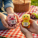 Two hands holding a pink pig and yellow duck plush toy with picnic setting in the background.