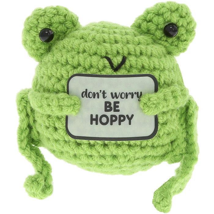 Green knitted frog toy with a sign saying 'don't worry BE HOPPY' on a white background