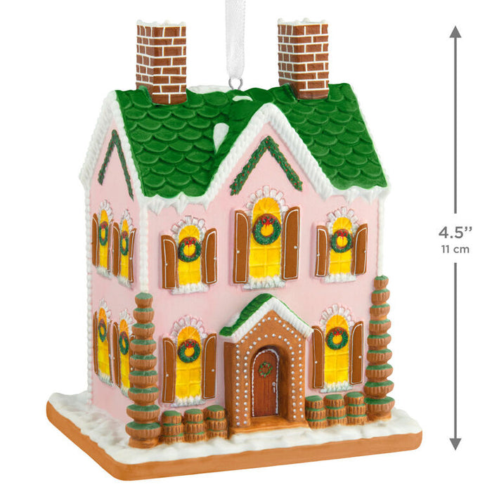 Decorative gingerbread house ornament with green roof and pink walls, featuring wreaths on windows.