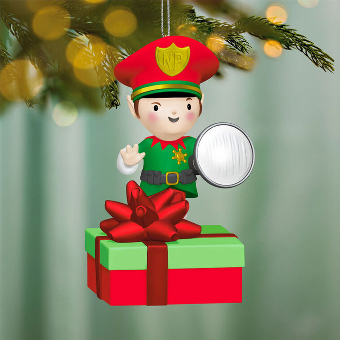 Christmas ornament of an elf on a present with a flashlight, hanging from a tree.