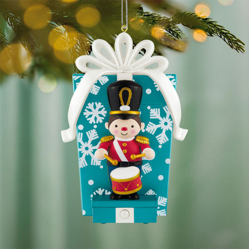 Christmas ornament of a drummer boy in a gift box with a white bow, against a blurred green background.