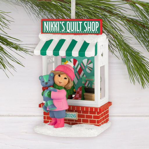 Christmas ornament of a girl with a teddy bear in front of 'Nikki's Quilt Shop' sign.