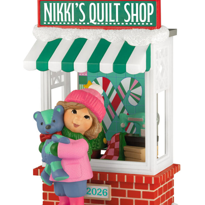 Decorative ornament of a girl with a teddy bear in front of 'Nikki's Quilt Shop'.