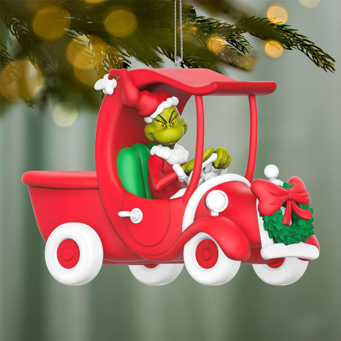 Christmas ornament of the Grinch in a red truck sleigh with a wreath, hanging against a blurred Christmas tree background.