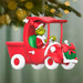 Christmas ornament of the Grinch in a red truck sleigh with a wreath, hanging against a blurred Christmas tree background.