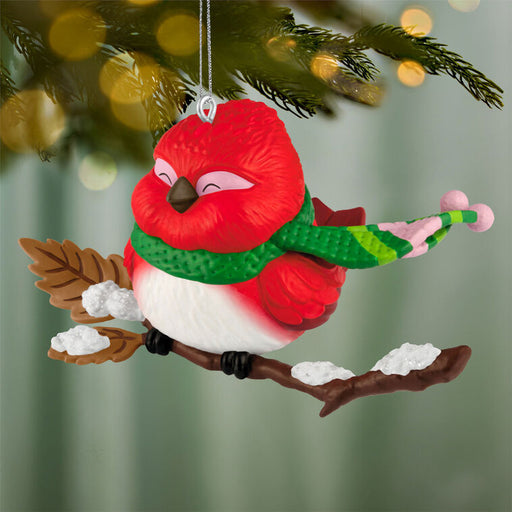 Red bird ornament with a green scarf on a branch against a blurred Christmas tree background.