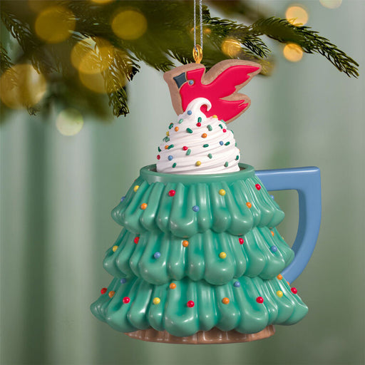 Decorative ornament shaped like a Christmas tree mug with a swirl of whipped cream and sparkles and topped with a cardinal cookie on a green background
