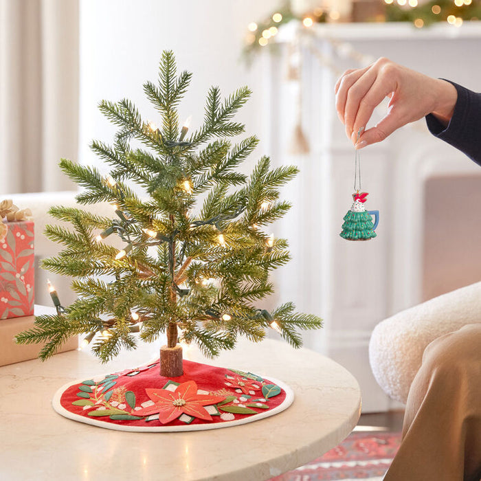Small decorated Christmas tree on a table with a person holding an ornament.