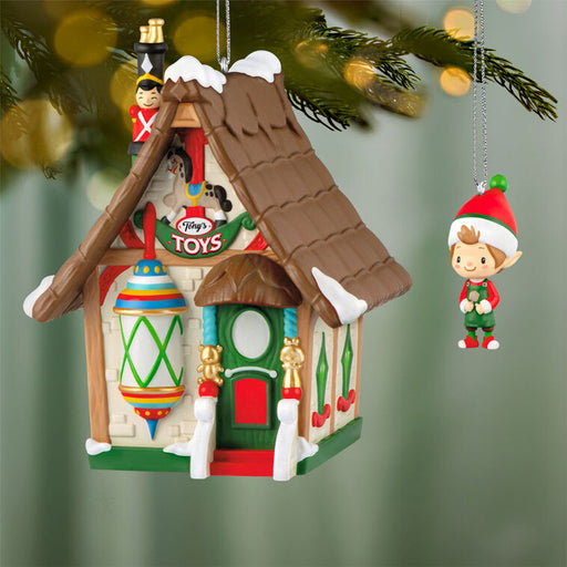 Decorative Christmas ornament of a toy store with an elf ornament hanging nearby, on a blurred green background.