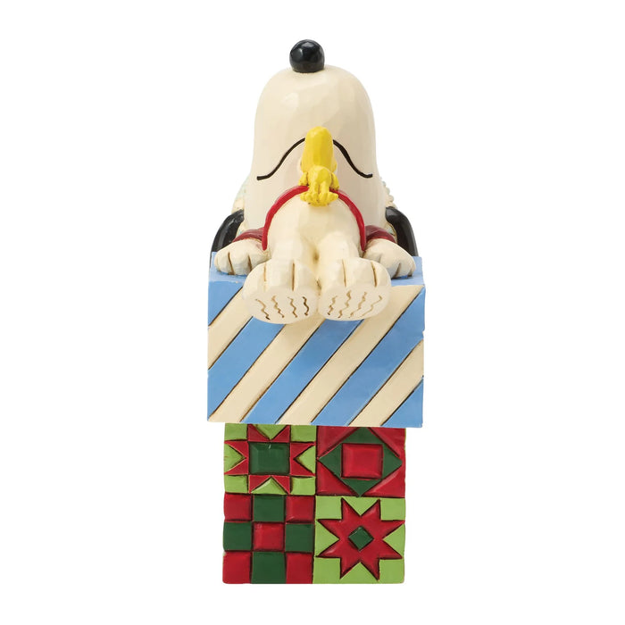 A collectible figurine of Snoopy peeking over a decorated Christmas gift box. The figurine features Snoopy's cream-colored face and paws, black ears, a yellow beak, and a red collar, emerging from a gift box with blue and cream diagonal stripes on top and a red, green, and blue quilted pattern on the bottom, all set against a plain white background.