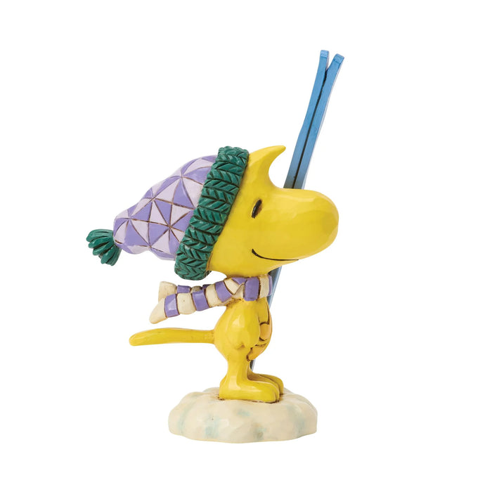 A collectible figurine of Snoopy wearing a purple and green geometric knit beanie and a matching striped knit scarf, holding a pair of blue skis on a small light-colored base against a white background.