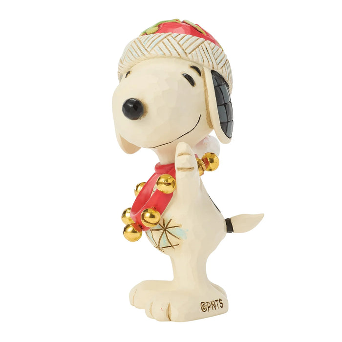 A collectible figurine of Snoopy wearing a holiday-themed outfit, featuring a cream-colored body, a red and gold patterned hat, a red scarf with gold bells, and a gold starburst design on his chest, set against a plain white background.