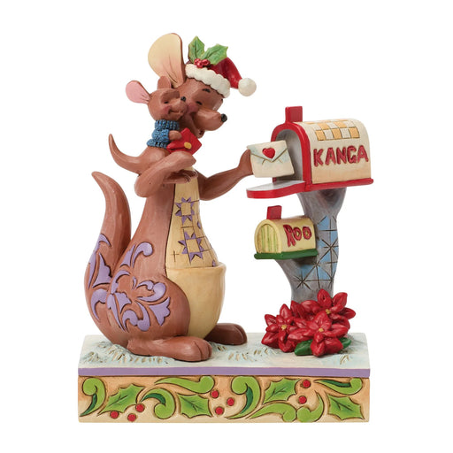 A Jim Shore figurine of a kangaroo wearing a Santa hat and scarf, kissing a mailman while standing next to a mailbox with a red envelope and poinsettias on a white background.