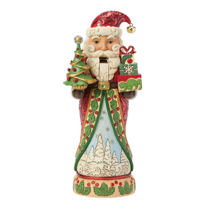 A Jim Shore Santa figurine holding a small decorated Christmas tree and a wrapped gift, set against a plain white background.