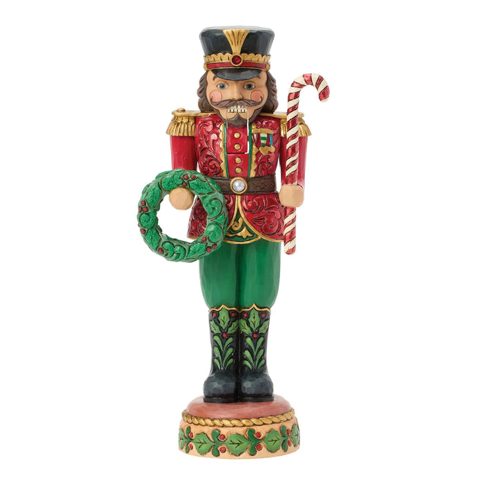 A festive Christmas nutcracker figurine standing on a decorated base, wearing a red coat, green pants, and holding a green wreath and a candy cane, isolated on a white background.