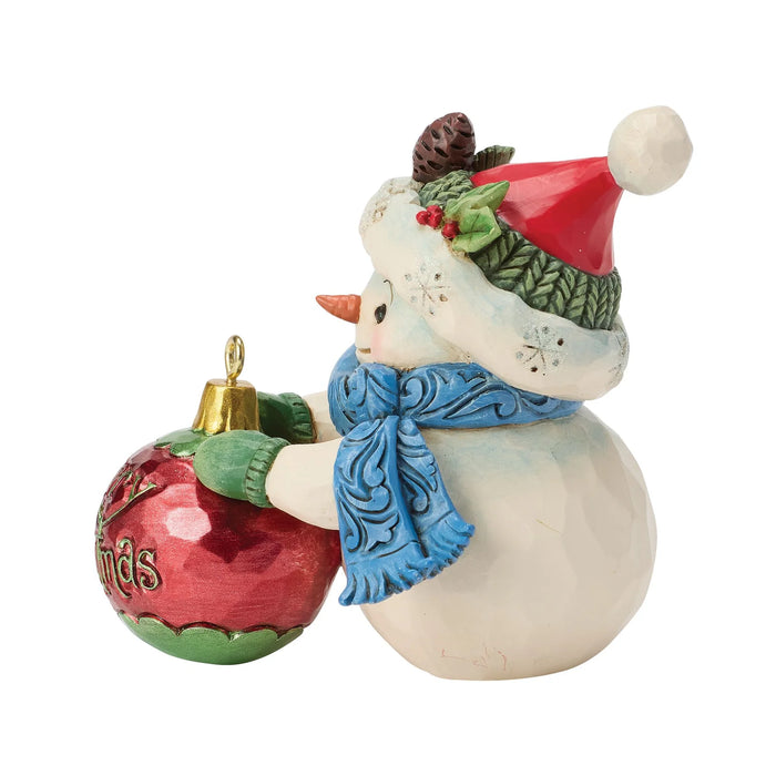 Snowman figurine with a hat and blue scarf holding a green and red ornament that says "Merry Christmas" on a white background